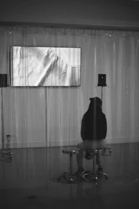 a black and white photo of a person sitting on a stool in front of a television